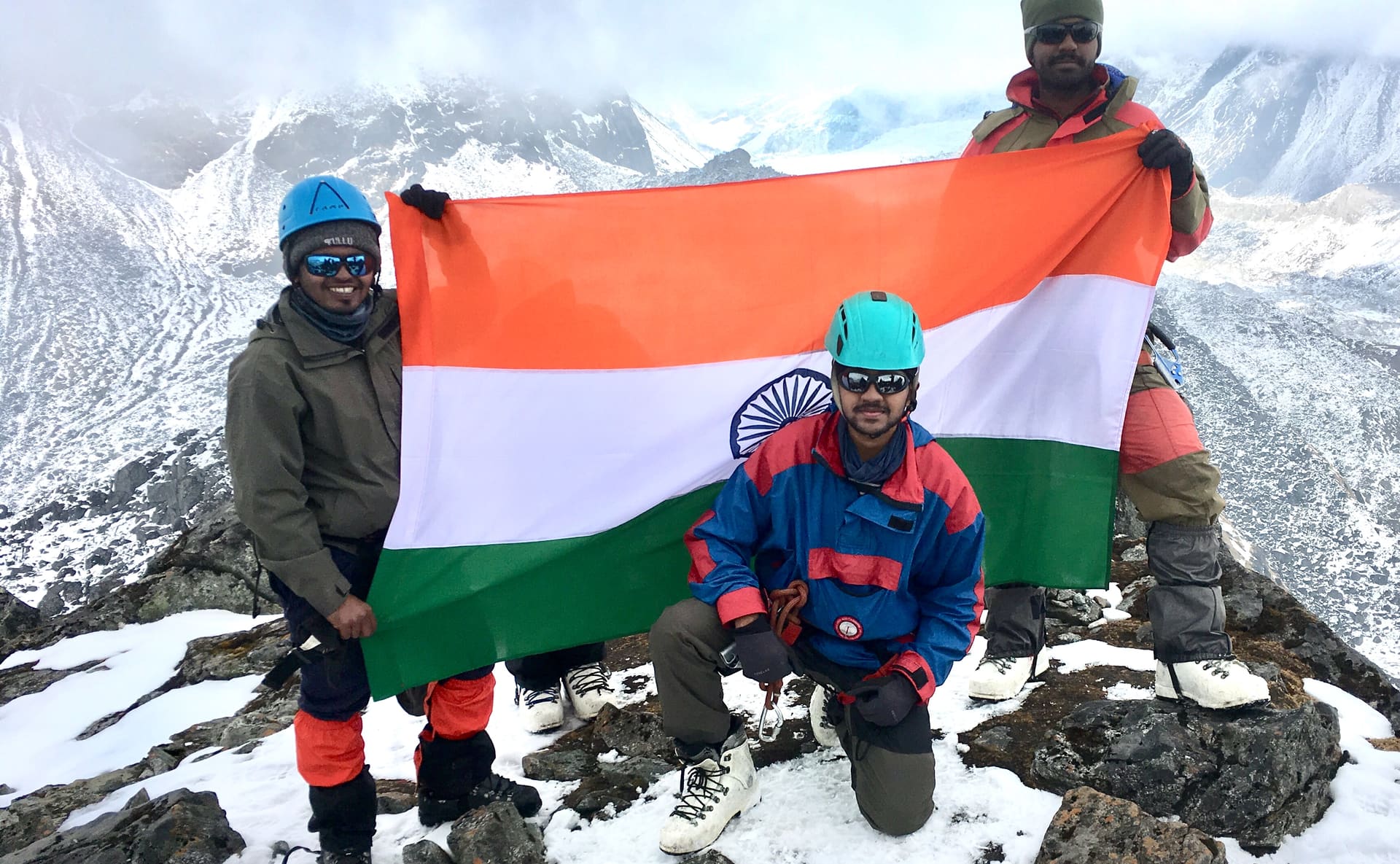 Three climbers in helmets and bright jackets hold the Indian flag on a rocky, snow-covered summit, with steep ice in the cloud behind them.
