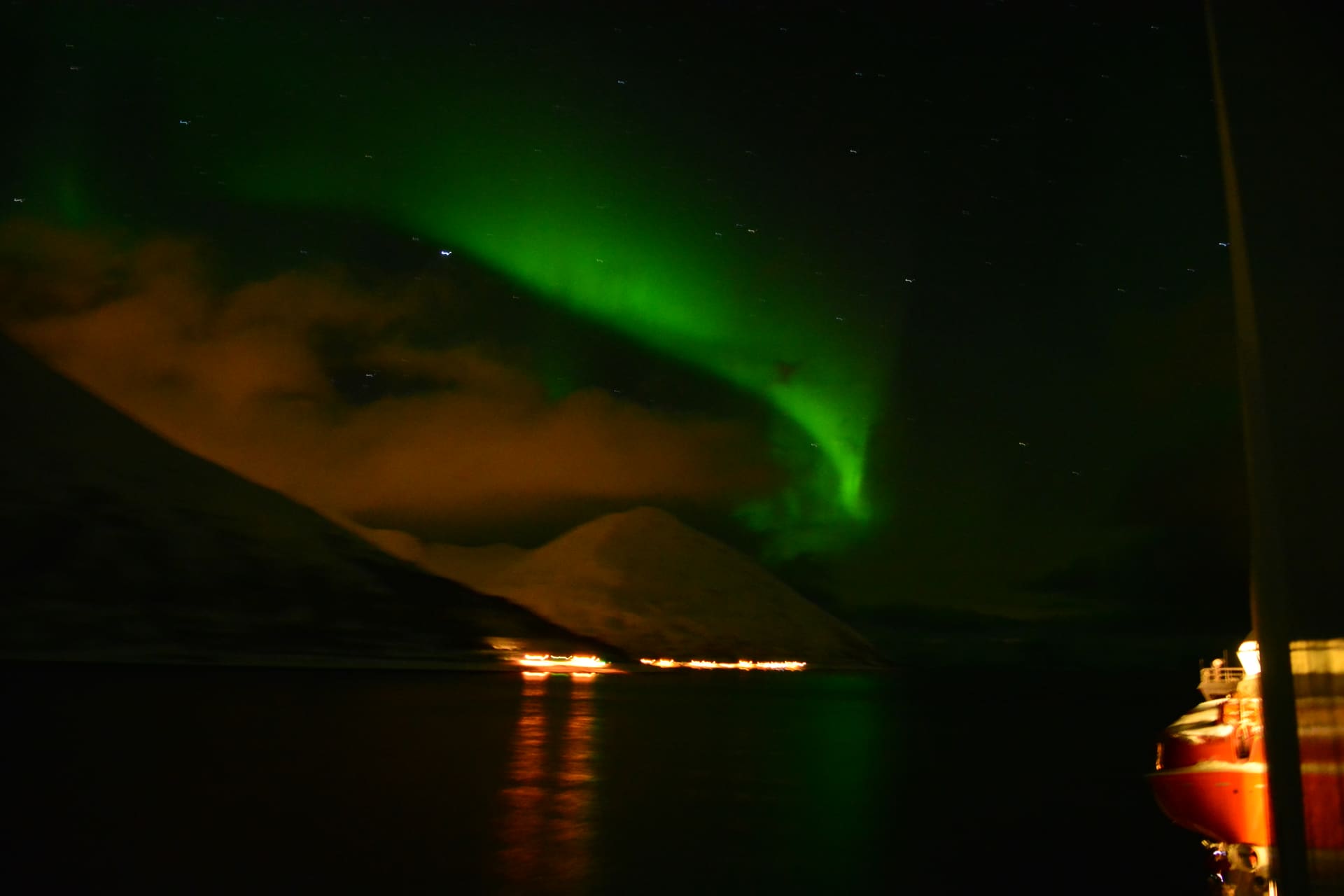 A green aurora arcs over a dark mountain and a fjord at night, with shore lights reflected in the water and part of a boat at the right edge.