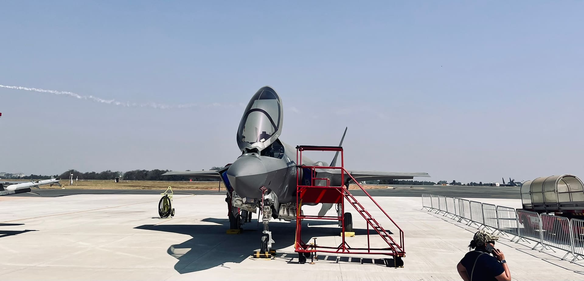 A grey fighter jet faces forward on a concrete apron, its canopy open, with a red boarding stair at the nose.