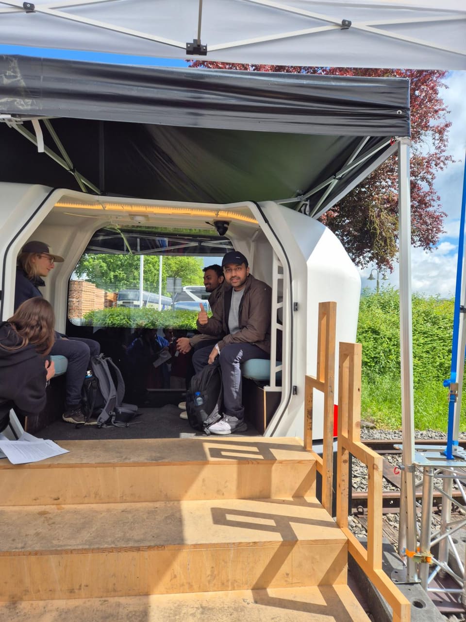 Arpan Ray sits in the open doorway of the white Monocab cabin and gives a thumbs-up, with wooden steps in front and other people seated inside.